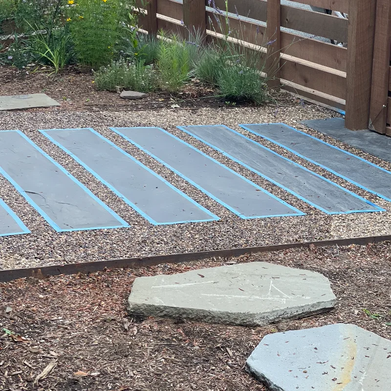 Intersecting pathways, one of rounded stones the other of sharp slate rectangular stone. Plants and a fence in the background.